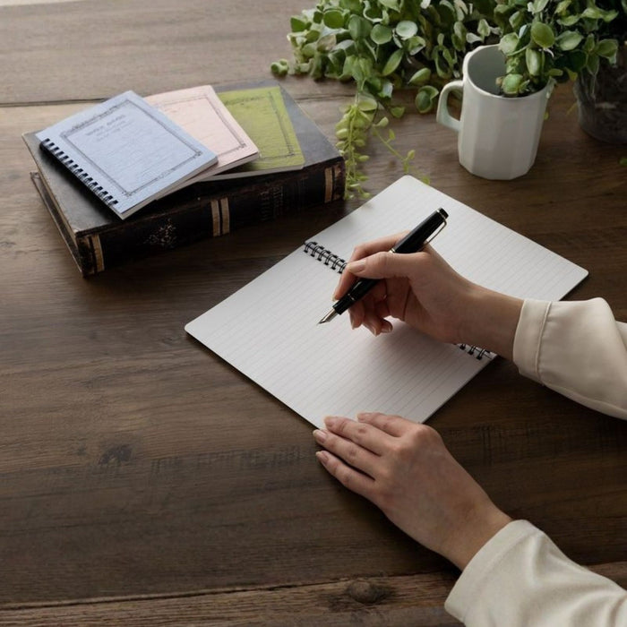 Person writing in an Apica CD Twin Ring Notebook on a desk with fountain pen friendly paper