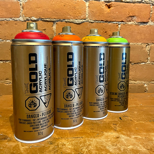 Line up of Montana Gold spray paint cans in red, orange, yellow, and green in front of brick wall.
